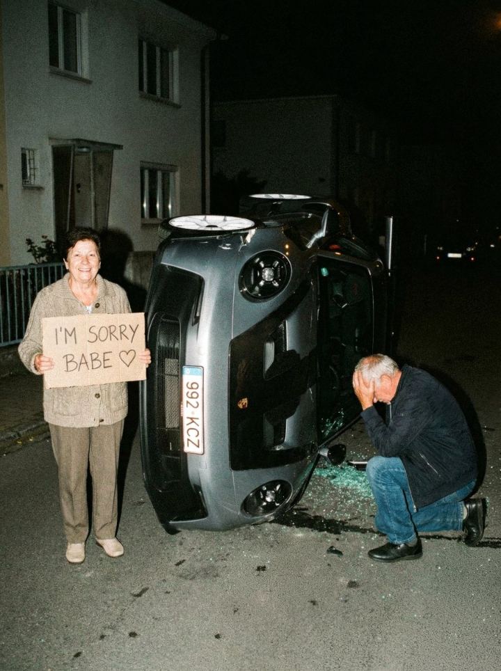 Older woman holding an “I'm sorry babe” sign beside an overturned Porsche at night